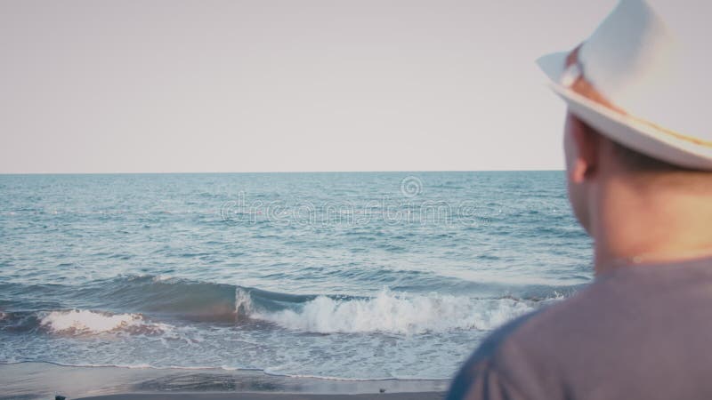 Un homme en chapeau debout sur le bord de mer regardant les vagues à l'envers banque de vidéos