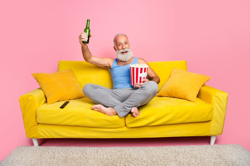Un homme âgé avec une barbe tenant de la bière en dégustant du pop-corn sur un canapé jaune dans un intérieur dynamique photo stock