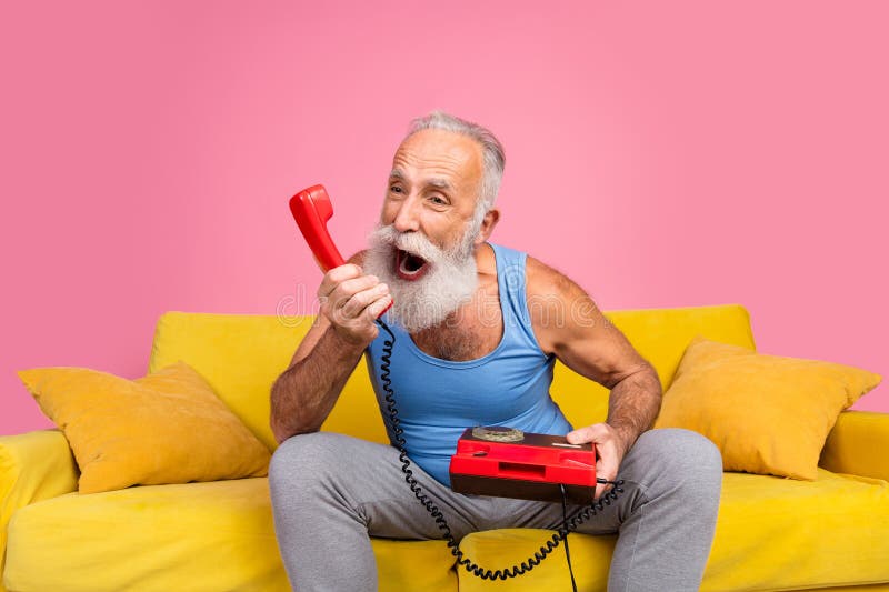 Un homme âgé gai avec une barbe assise sur un canapé jaune tenant un téléphone rouge vintage image libre de droits