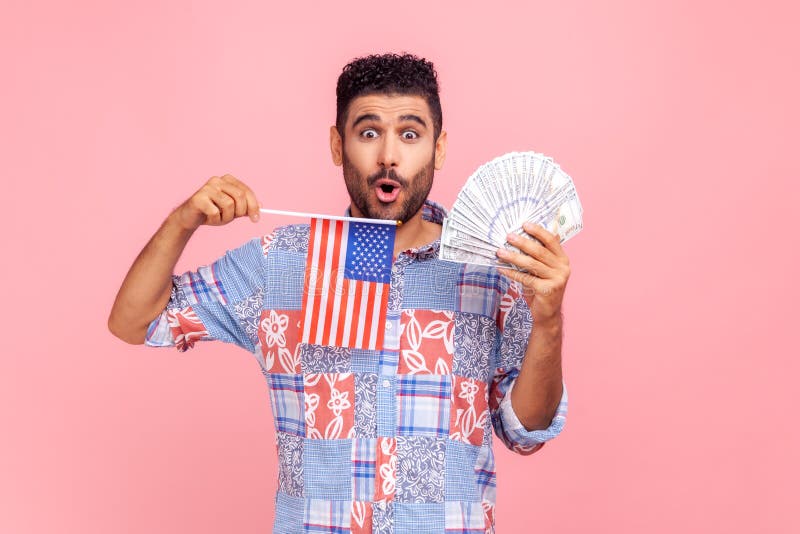 Un homme étonnant et attrayant avec barbe et cheveux foncés tenant un drapeau américain et des billets de dollars regardant la cam photographie stock