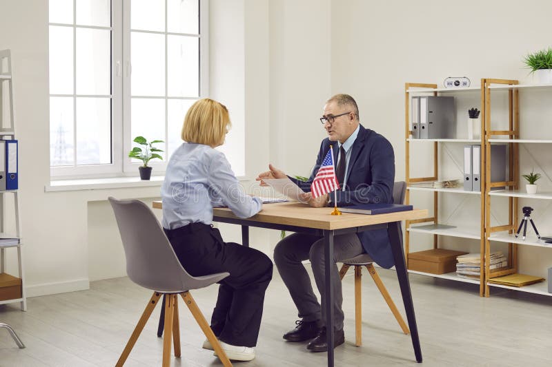 Government authority or United States of America embassy consul discusses visa application with customer at table in office. Male Immigration Services agency worker meeting with woman who moved to USA. Government authority or United States of America embassy consul discusses visa application with customer at table in office. Male Immigration Services agency worker meeting with woman who moved to USA