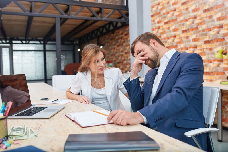 Un impiegato di concetto maschio copre il suo fronte della frustrazione dai problemi, il suo collega è le comodità lui immagine stock Un impiegato di concetto maschio copre il suo fronte della frustrazione dai problemi, il suo collega è le comodità lui immagine stock