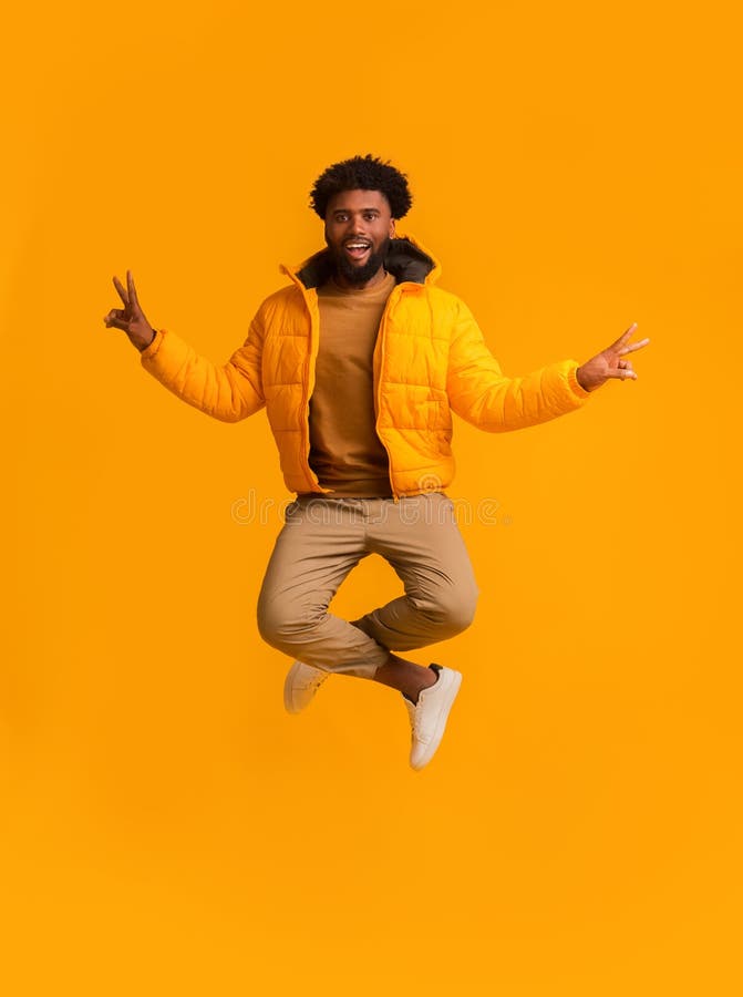 Peaceful winter african guy demonstrating peace symbol while jumping, orange background. Peaceful winter african guy demonstrating peace symbol while jumping, orange background