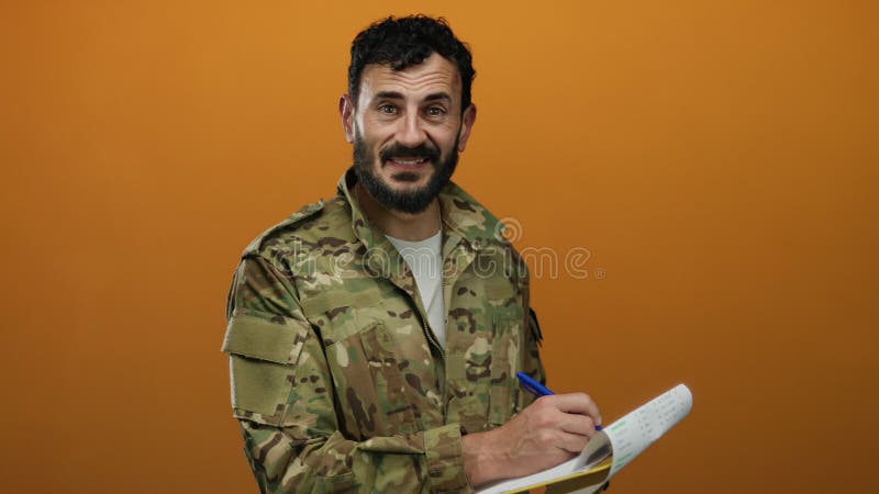 Un ispanico con la barba in uniforme militare scrive su un taccuino con un vivido sfondo arancione fotografia stock libera da diritti