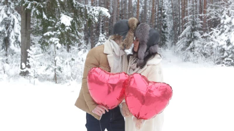 Un jeune couple heureux se tient en face et s'embrasse dans une forêt d'hiver enneigée tenant en forme de coeur rouge banque de vidéos