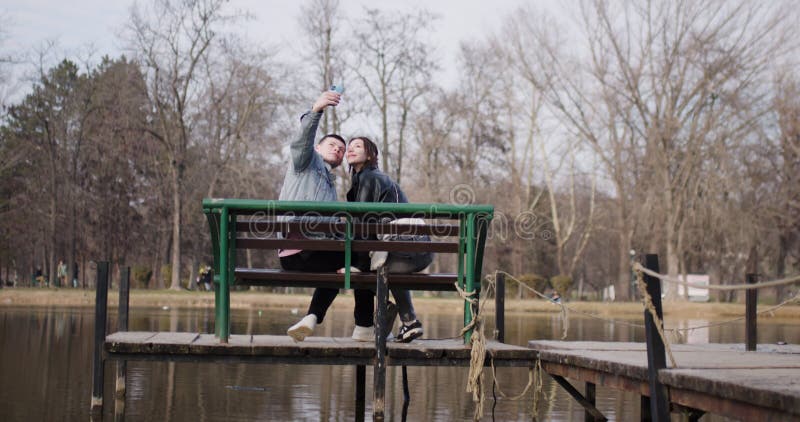 Un jeune couple prend des selfies sur un banc de parc surplombant un lac clips vidéos