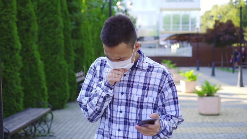 Un jeune homme asiatique avec un masque médical sur le visage toussant et utilisant un smartphone