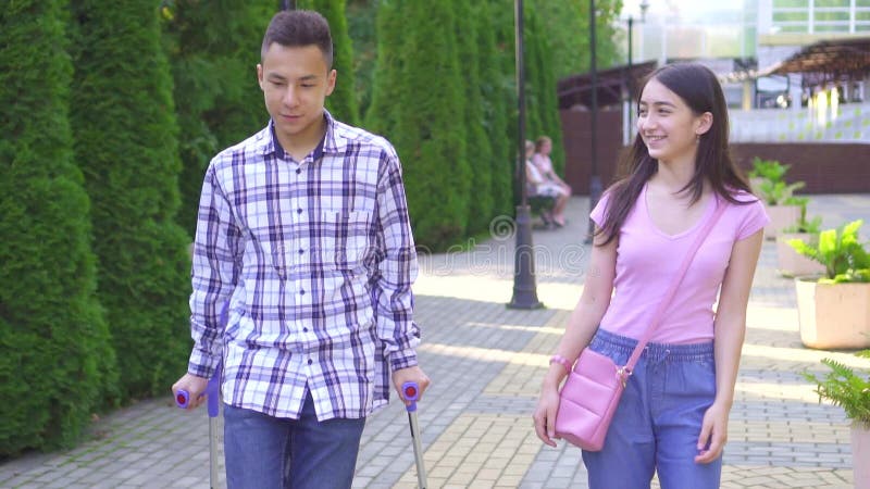 Un jeune homme asiatique qui a une jambe cassée sur des béquilles communique avec une jeune femme asiatique dans le parc, près de