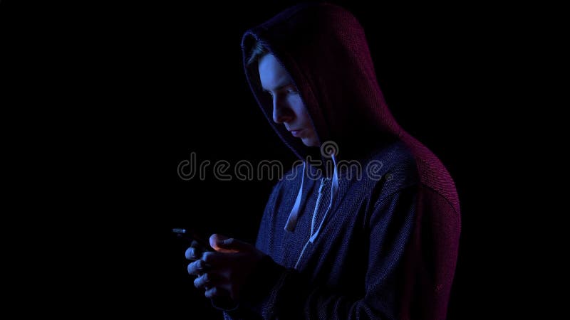 A young man in a hood with a phone in his hands. Hacker makes a hack through the phone. Blue and red light falls on a person on a black background. 4k. A young man in a hood with a phone in his hands. Hacker makes a hack through the phone. Blue and red light falls on a person on a black background. 4k