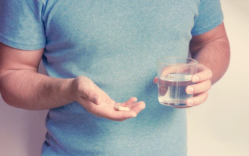 Un jeune homme dans des t-shorts bleus tient un groupe de capsules avec la médecine ou de vitamines dans ses mains et un verre de images libres de droits Un jeune homme dans des t-shorts bleus tient un groupe de capsules avec la médecine ou de vitamines dans ses mains et un verre de images libres de droits