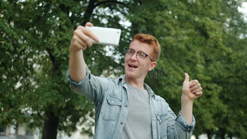 Un jeune homme joyeux qui prend le selfie avec un appareil photo de son portable à l'extérieur du parc banque de vidéos