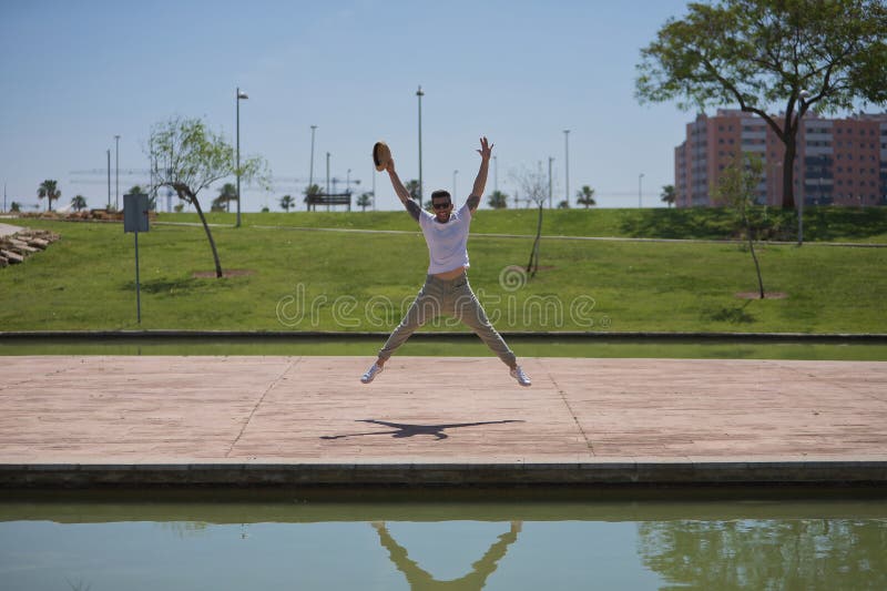 Un joven atractivo e hispano vestido con una camiseta blanca y un sombrero saltando al aire muy feliz en un parque exterior. conce imágenes de archivo libres de regalías