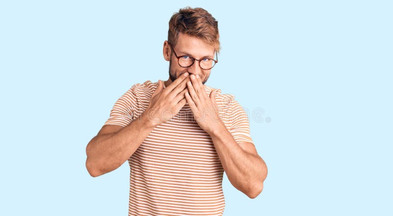 Un joven caucásico con ropa informal y gafas riéndose y riéndose avergonzado mientras cuida la boca con chismes en las manos y fotos de archivo libres de regalías