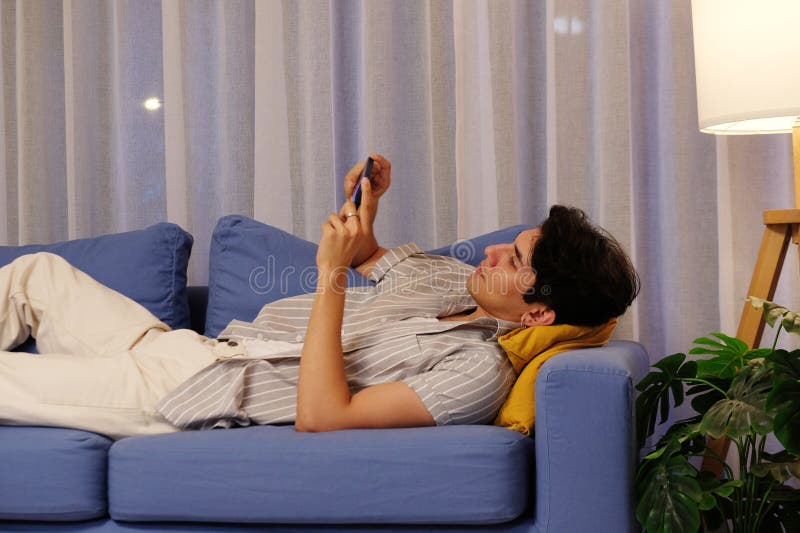 Un joven feliz usando un teléfono móvil mientras descansa en un sofá por la noche, relajándose en casa imagen de archivo libre de regalías