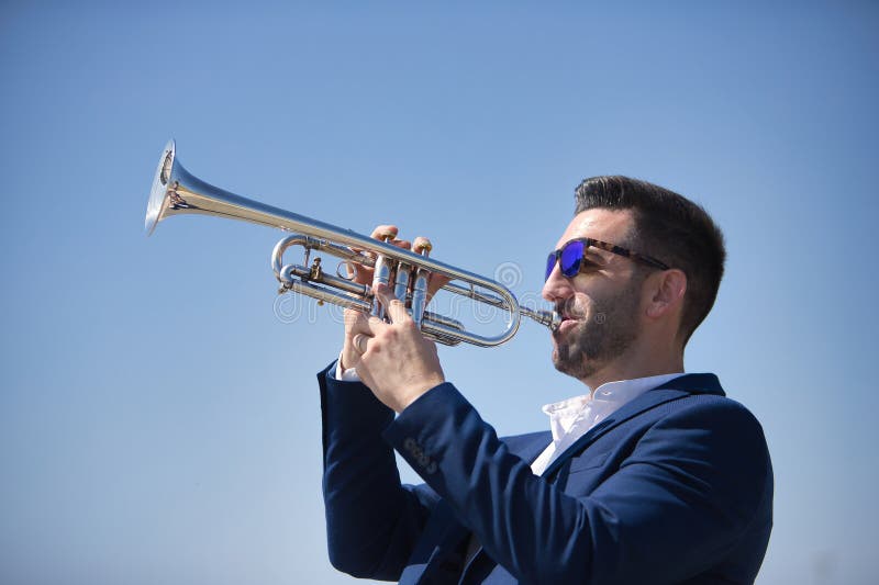 Un joven hispano con una chaqueta y gafas de sol jugando una trompeta de plata al aire libre. instrumentos de música de conceptos imágenes de archivo libres de regalías