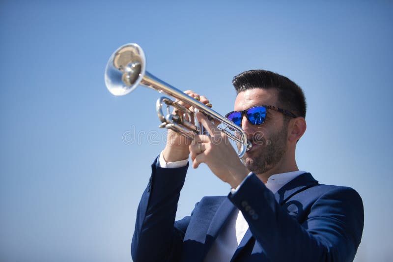 Un joven hispano con una chaqueta y gafas de sol jugando una trompeta de plata al aire libre. instrumentos de música de conceptos fotografía de archivo libre de regalías