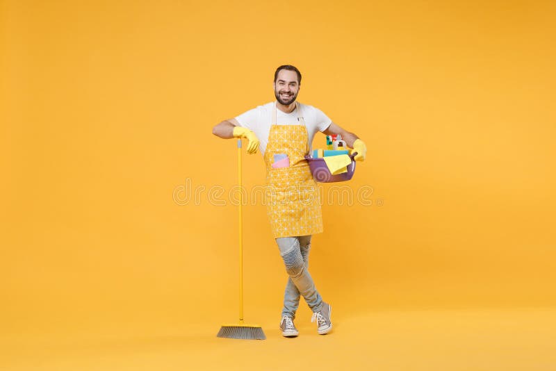 Un joven hombre sonriente ama de casa en guantes de delantal sostienen botellas detergentes lavadoras escoba de lavadoras de cuenc foto de archivo libre de regalías