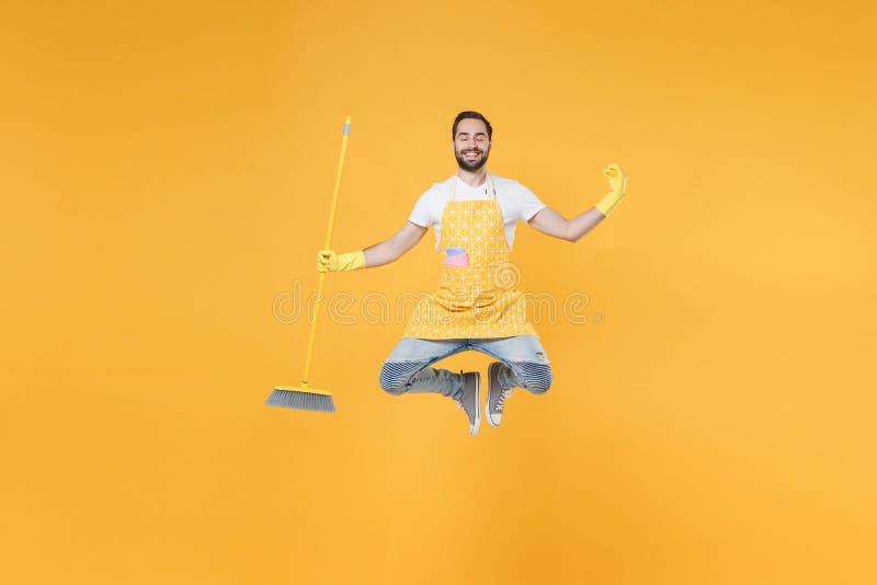 Un joven hombre sonriente esposo de la casa con guantes de goma de delantal sostiene escoba mientras realiza tareas domésticas ais fotografía de archivo libre de regalías