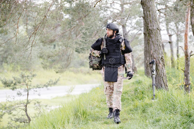 Un hombre con uniforme militar y chaleco antibalas se sienta en el bosque  cerca de un detector de metales.