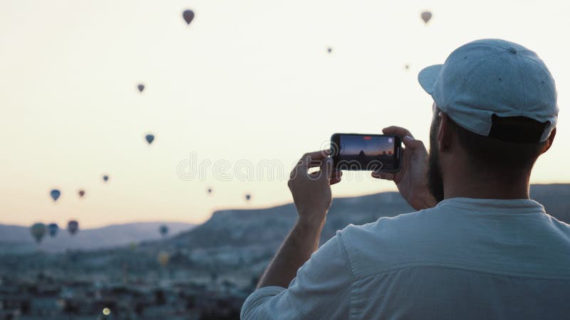 Un joven turista emocionado tomando un video con un smartphone con una hermosa vista de globos de aire caliente. nevada de cappado