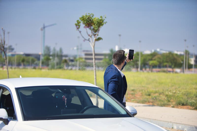 Un joven y exitoso hombre hispano con chaqueta y gafas de sol tomándose un selfie con su celular al lado de su lujo blanco fotografía de archivo libre de regalías