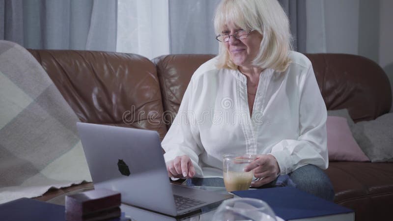 Un jubilado caucásico positivo sentado en un sofá con una taza de café y mirando la pantalla de un portátil Mujer rubia de alto n almacen de metraje de vídeo