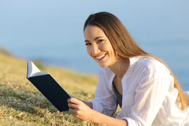 Happy reader holding a paper book lying on the grass looks at you. Happy reader holding a paper book lying on the grass looks at you