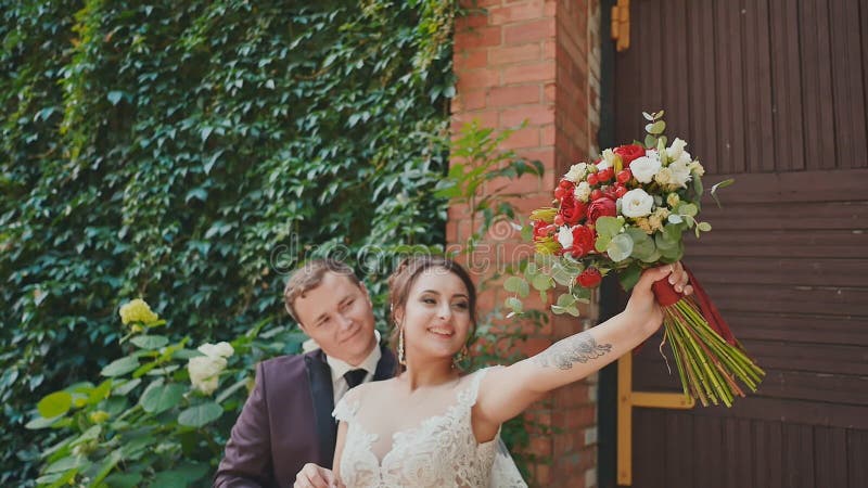 Un marié élégant et une belle jeune mariée avec un bouquet lumineux sur le fond d'un mur de briques avec les branches croissantes