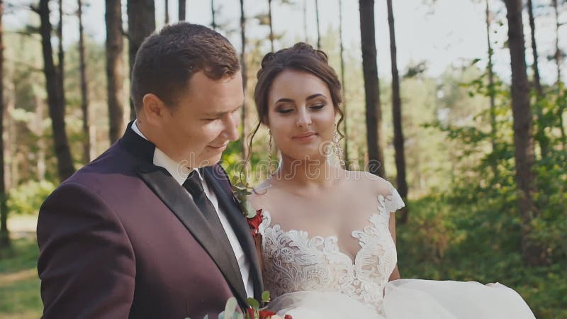 Un marié élégant et une jeune mariée avec du charme avec un bouquet lumineux, marchant le long d'une traînée de forêt parmi les p