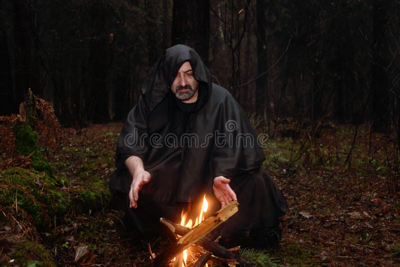 A black-robed monk sits by a fire in the forest and looks into a divination ball. Mysticism and magic, sorcerers and witches. A black-robed monk sits by a fire in the forest and looks into a divination ball. Mysticism and magic, sorcerers and witches