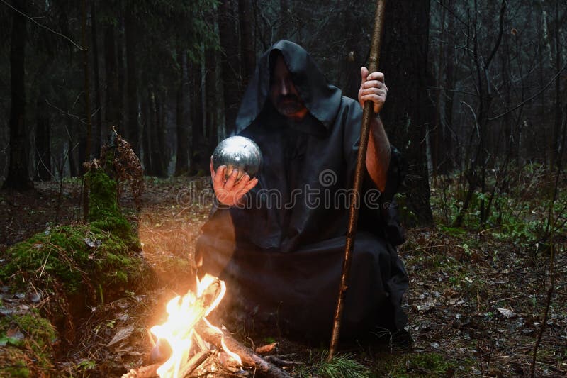A black-robed monk sits by a fire in the forest and looks into a divination ball. Mysticism and magic, sorcerers and witches. A black-robed monk sits by a fire in the forest and looks into a divination ball. Mysticism and magic, sorcerers and witches