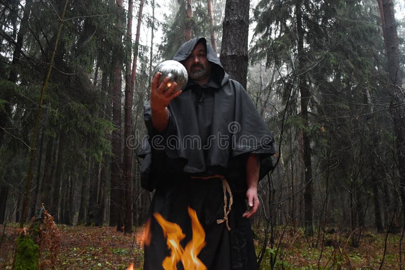 A black-robed monk sits by a fire in the forest and looks into a divination ball. Mysticism and magic, sorcerers and witches. A black-robed monk sits by a fire in the forest and looks into a divination ball. Mysticism and magic, sorcerers and witches