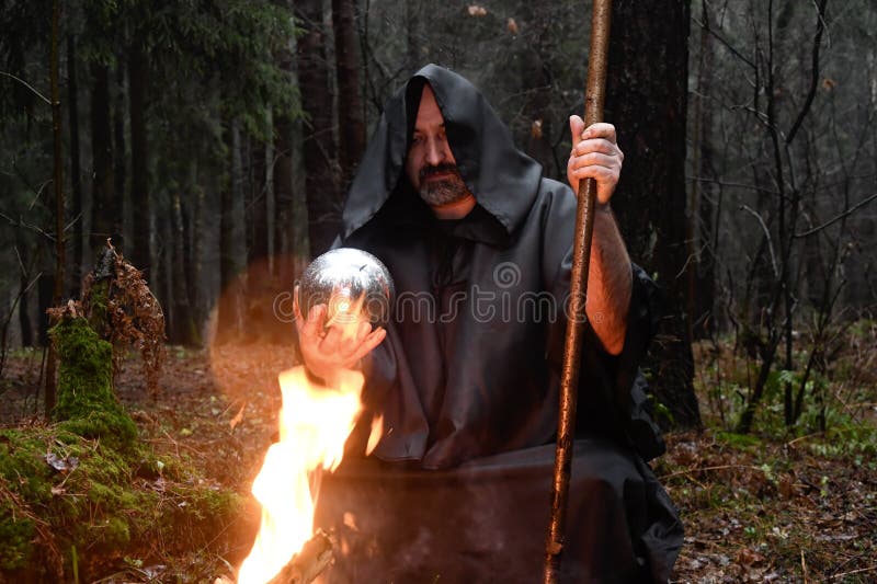 A black-robed monk sits by a fire in the forest and looks into a divination ball. Mysticism and magic, sorcerers and witches. A black-robed monk sits by a fire in the forest and looks into a divination ball. Mysticism and magic, sorcerers and witches
