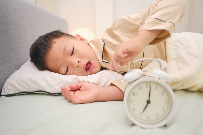 Un niño adorable y pequeño tendido en la cama y parando de apagar el despertador en el dormitorio en casa fotos de archivo