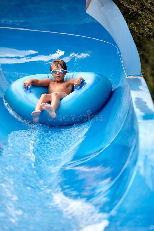 Un niño alegre desciende sobre un círculo inflable desde el tobogán de agua en el parque acuático los niños atracciones en foto de archivo