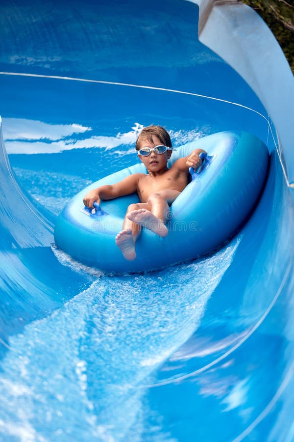 Un niño alegre nadando gafas se desliza por el tobogán de agua azul en un bote inflable en el parque acuático salpican agua imágenes de archivo libres de regalías
