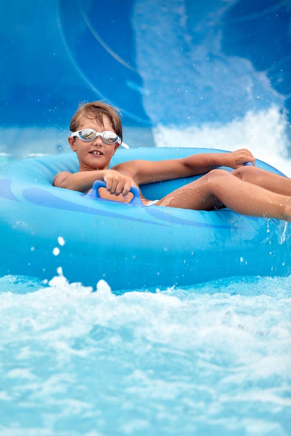 Un niño alegre nadando gafas se desliza por el tobogán de agua azul en un bote inflable en el parque acuático salpican agua fotos de archivo libres de regalías