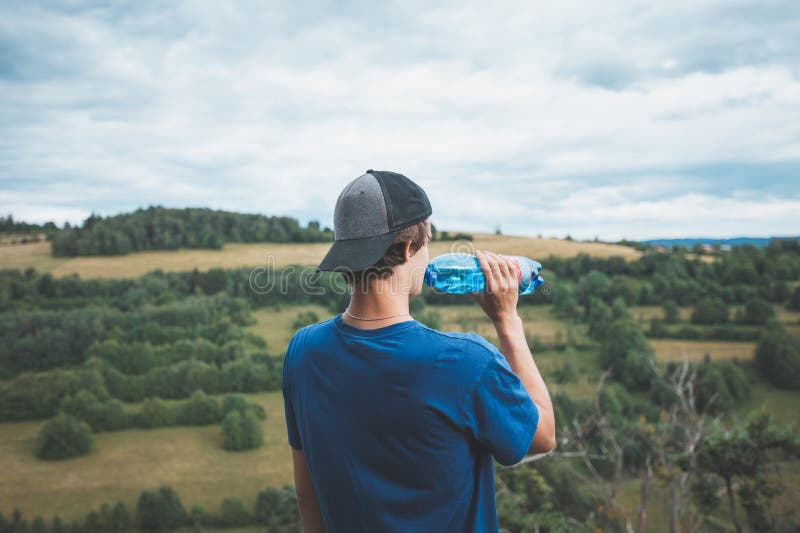 Un niño con camisa y gorra azul disfruta de una bebida refrescante de una botella de agua mientras admira los extensos campos verd foto de archivo libre de regalías