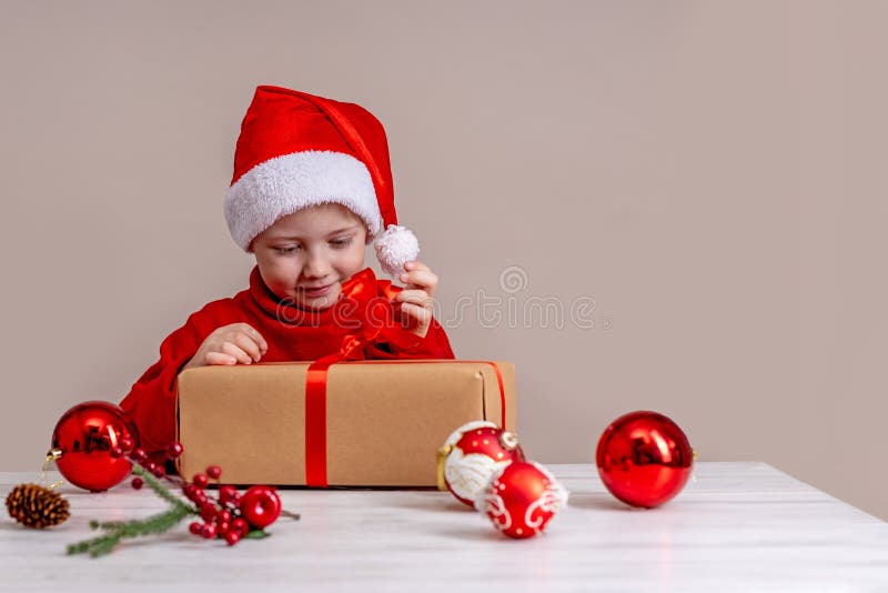 Un niño con traje de santa claus mirando regalos de navidad. retrato de cierre sobre fondo beige foto de archivo libre de regalías