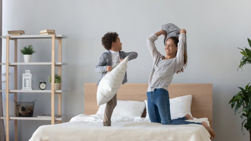Overjoyed cute little mixed race preschool boy attacking laughing mommy with pillow on bed. Happy young single women playing fighting having fun with energetic small son in modern bedroom at home. Overjoyed cute little mixed race preschool boy attacking laughing mommy with pillow on bed. Happy young single women playing fighting having fun with energetic small son in modern bedroom at home.