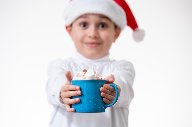 Un niño en un sombrero rojo de santa sonríe y muestra a la cámara una taza azul con los malvaviscos. Concepto de Navidad imagen de archivo libre de regalías