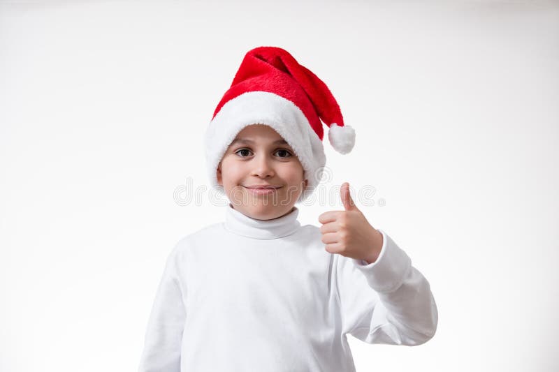 Un niño en un sombrero rojo de santa sonríe y muestra su dedo pulgar. Concepto de Navidad fotografía de archivo libre de regalías
