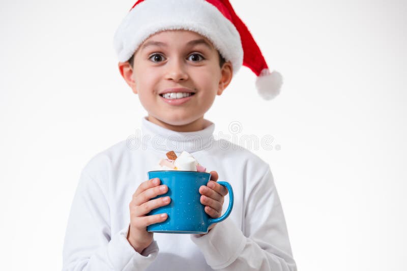 Un niño en un sombrero rojo de santa sonríe y sostiene una taza de malvaviscos. Concepto de Navidad imagenes de archivo