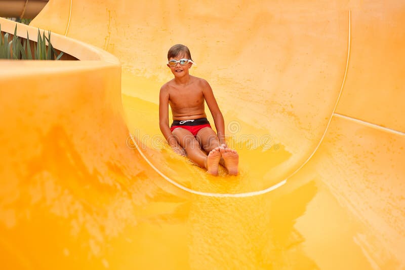Un niño pequeño con gafas se desliza por un tobogán amarillo en el parque acuático un ánimo alegre para los niños vacaciones fotos de archivo libres de regalías