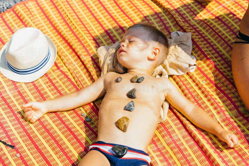 Un niño pequeño es el tomar el sol de mentira en una estera anaranjada con las piedras en su estómago fotografía de archivo libre de regalías Un niño pequeño es el tomar el sol de mentira en una estera anaranjada con las piedras en su estómago fotografía de archivo libre de regalías