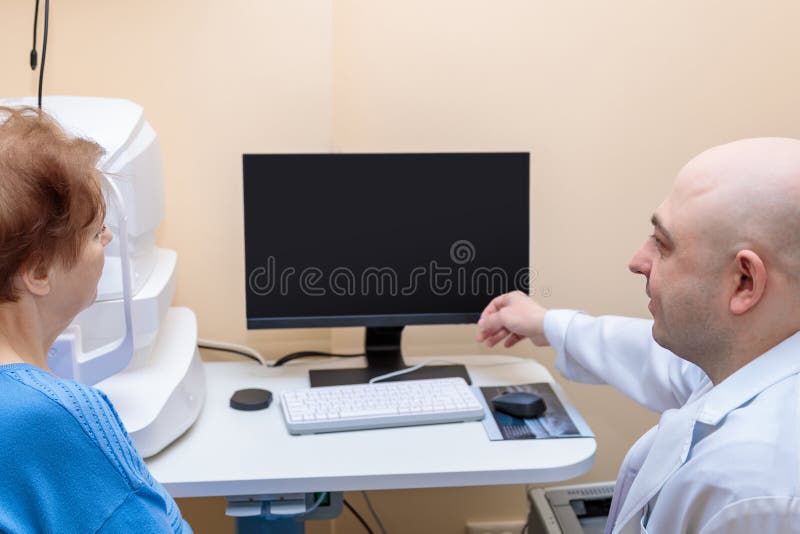 Un oftalmologo maschio spiega i risultati di un esame oculare effettuato da una donna adulta che mostra un disegno su un computer fotografia stock