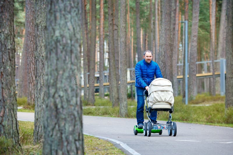 Un padre joven en tablero de la vespa del girocompás con un cochecito camina a través del parque del otoño imagen de archivo Un padre joven en tablero de la vespa del girocompás con un cochecito camina a través del parque del otoño imagen de archivo