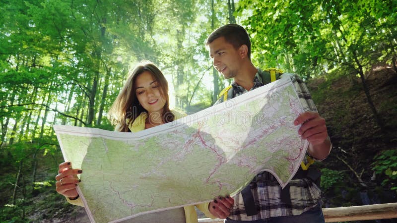 Un par joven de los turistas que miran un mapa Se colocan en los rayos del sol en el bosque cerca de la cascada