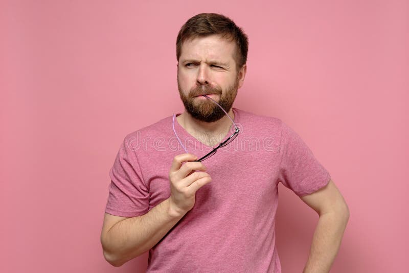 Pensive Caucasian bearded man suspects someone, he holds glasses at mouth and looks with a squint. Isolated on a pink background. Pensive Caucasian bearded man suspects someone, he holds glasses at mouth and looks with a squint. Isolated on a pink background.