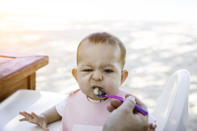 Un pequeño bebé está enojado y no quiere comer con una cuchara La niña pequeña se sienta en una silla en la playa en el día imagen de archivo Un pequeño bebé está enojado y no quiere comer con una cuchara La niña pequeña se sienta en una silla en la playa en el día imagen de archivo